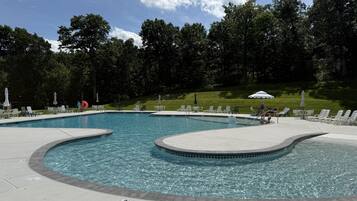 A swimming pool with multiple lanes, surrounded by lounge chairs and umbrellas, set against a backdrop of trees and a grassy slope.