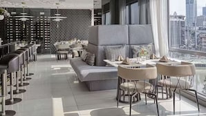 Un espace lounge moderne comprenant un canapé, des tabourets de bar et des tables, et offrant une vue sur la ville grâce à de grandes fenêtres.