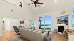 A spacious living room with a large window overlooking the ocean, a fireplace, and a ceiling fan. - Peaceful Coastal Estate | Beach Access + Sunrises (Palm Coast)