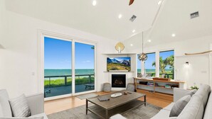 A spacious living room with a large sliding glass door leading to a balcony with a view of the ocean. There is a fireplace, a television, and a dining area with a table and chairs. - Peaceful Coastal Estate | Beach Access + Sunrises (Palm Coast)