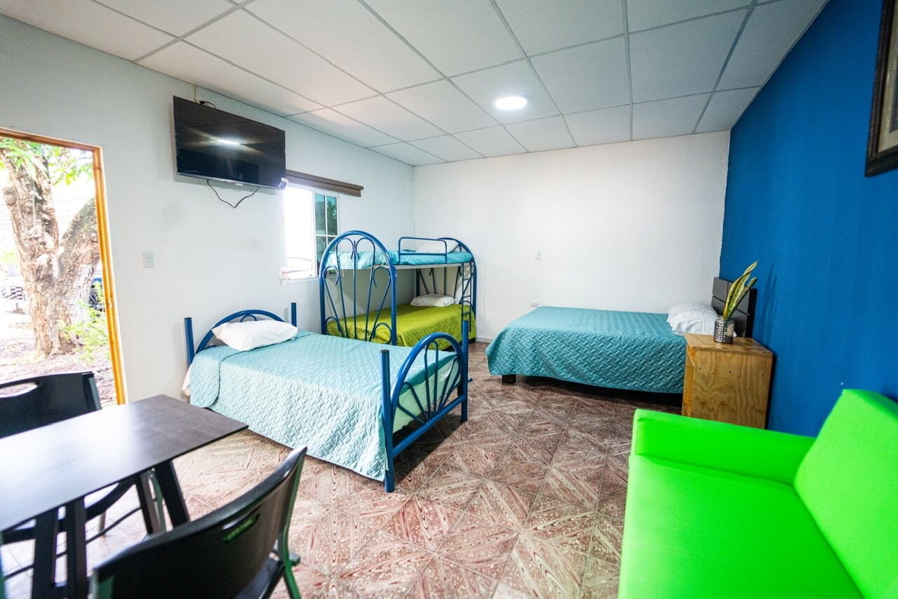 A dormitory room with bunk beds, a TV mounted on the wall, a small table, and a green chair.