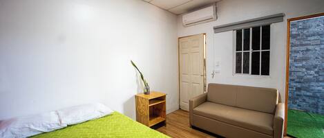 Quarto com uma cama, um sofá, uma mesa de cabeceira de madeira com uma planta, uma porta e uma janela.