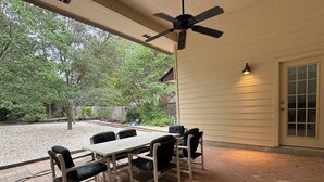 Un patio exterior con mesa y sillas, un ventilador de techo y vista a zona boscosa.