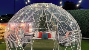 A geodesic dome structure with a bed and seating area inside, surrounded by artificial grass and illuminated by string lights.