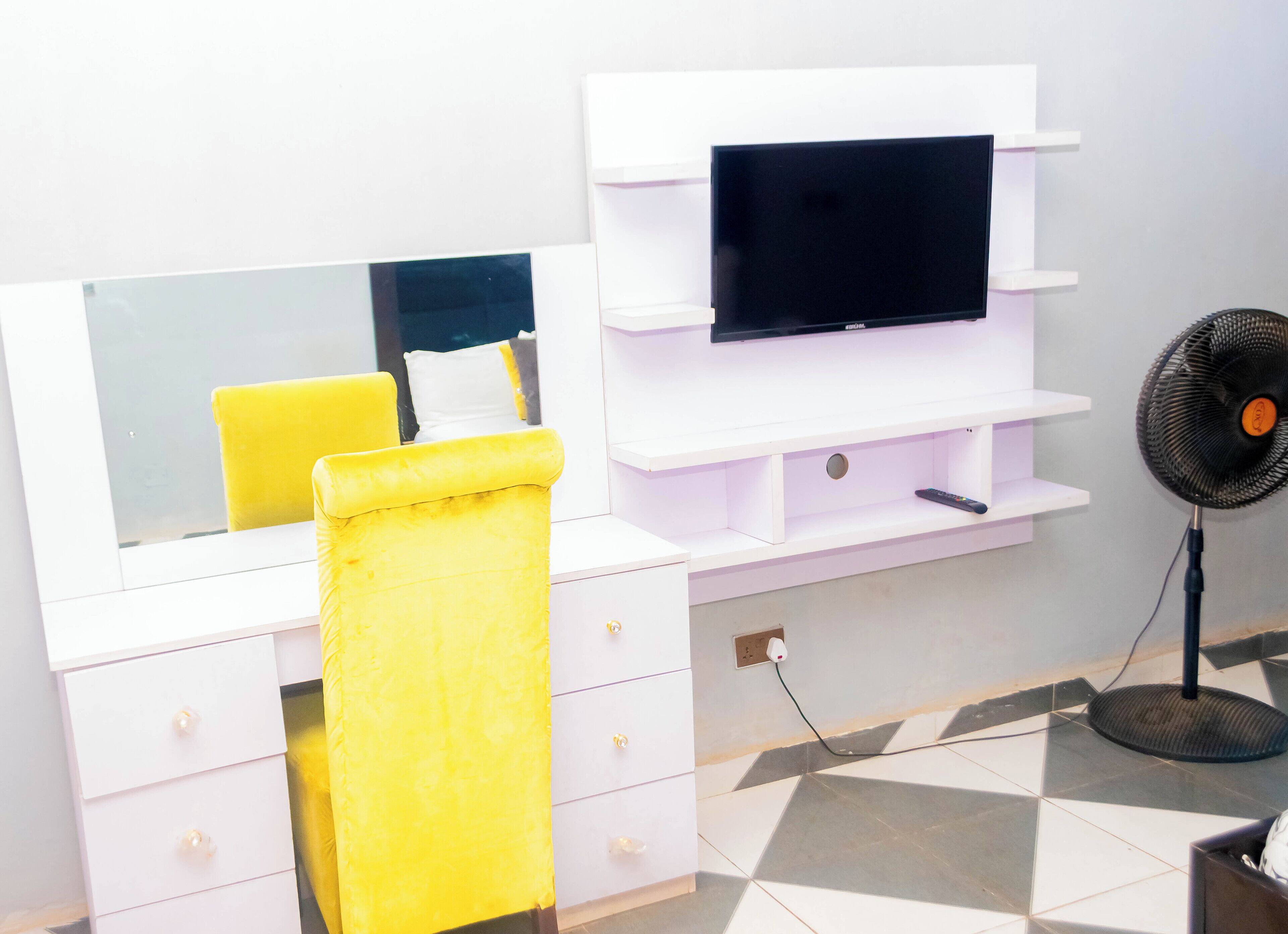 Una habitación con un tocador blanco, una silla amarilla, un televisor de pantalla plana montado en un estante blanco y un ventilador de pie.