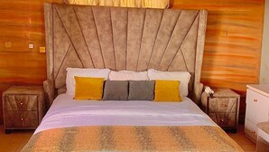 A bedroom with a large bed, wooden panel walls, a tufted headboard, and a nightstand with a lamp. - MC RICA HOTEL (Ikorodu)