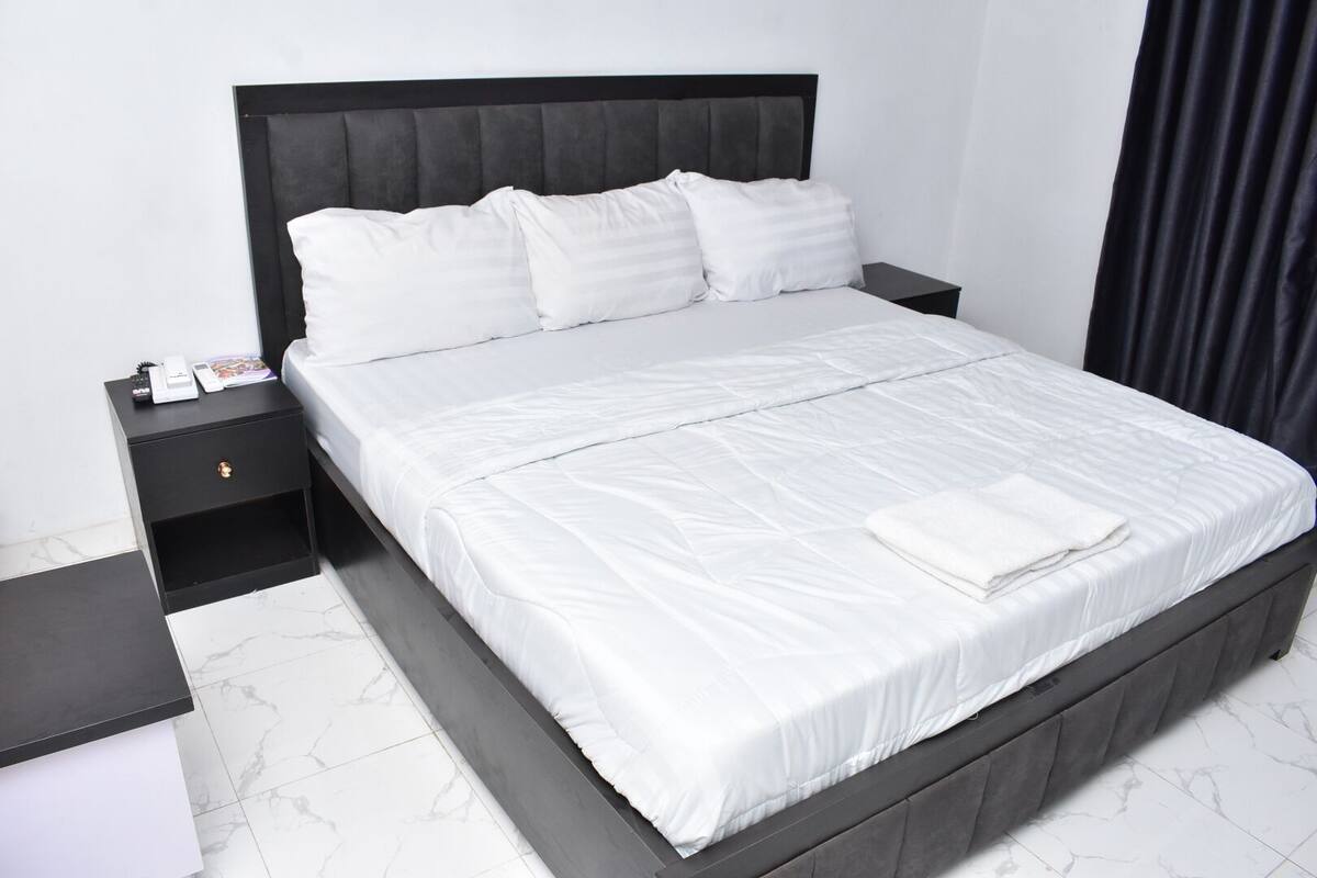 A neatly made bed with white linens, a black headboard, and a small bedside table with a drawer.