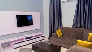 A modern living room with a flat-screen TV mounted on a white wall, a brown sofa with yellow cushions, a coffee table, and a tiled floor. - MC RICA HOTEL (Ikorodu)