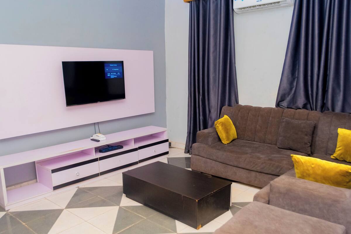 A modern living room with a flat-screen TV mounted on a white wall, a brown sofa with yellow cushions, a coffee table, and a tiled floor.