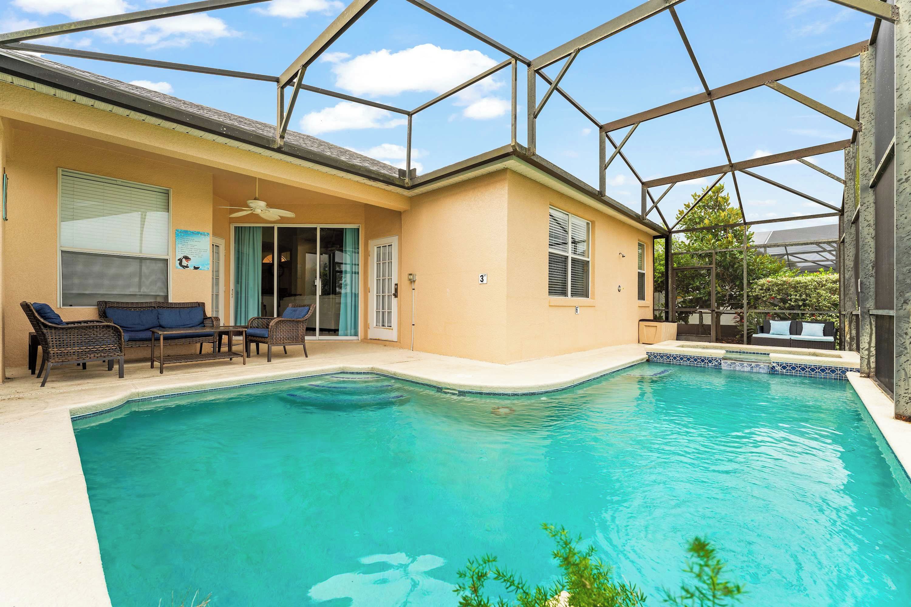 A swimming pool with a glass enclosure, a patio with wicker furniture, and a door leading to a covered area.
