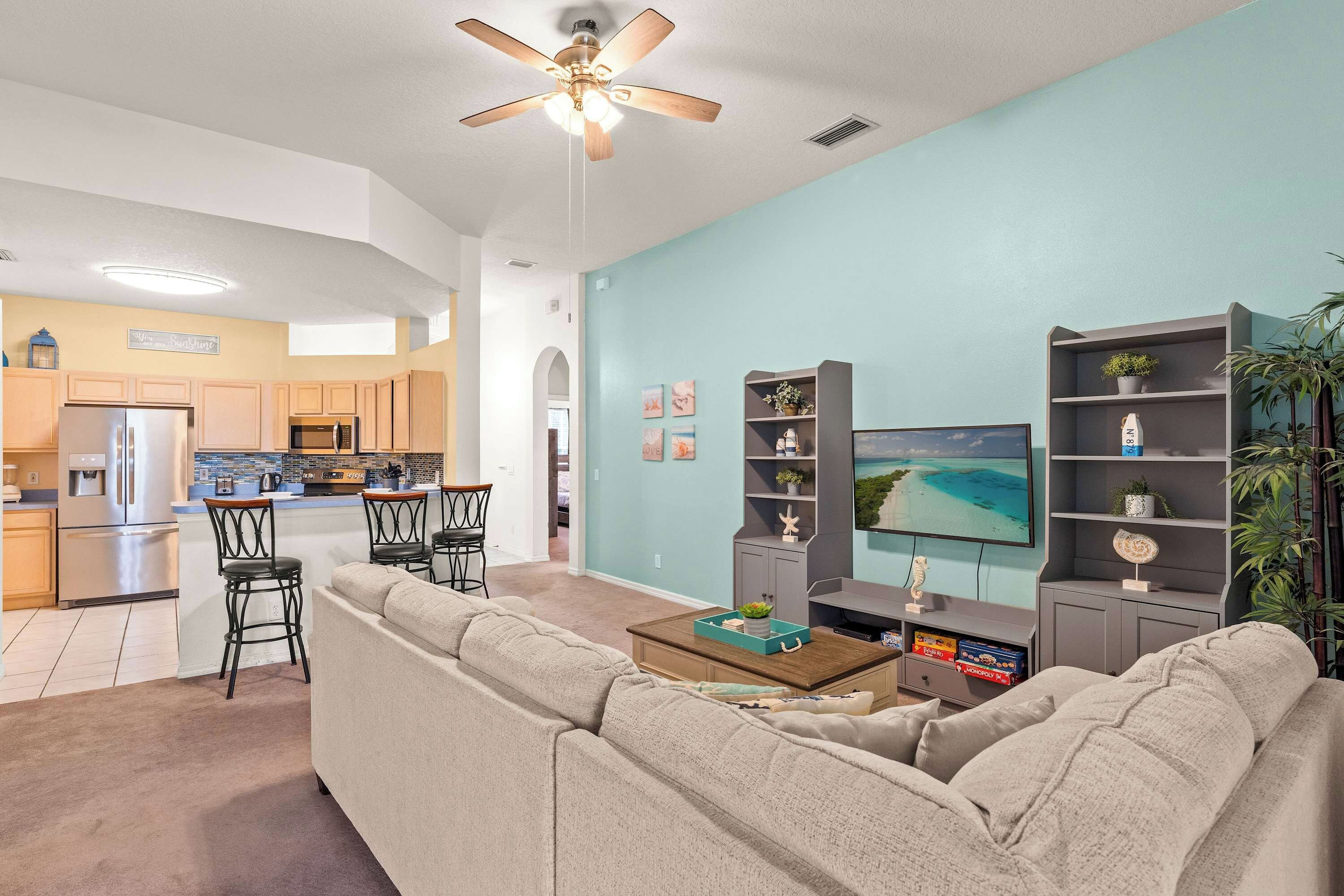 A modern living room with a sofa, coffee table, and TV. A kitchen with a refrigerator and microwave is visible in the background.