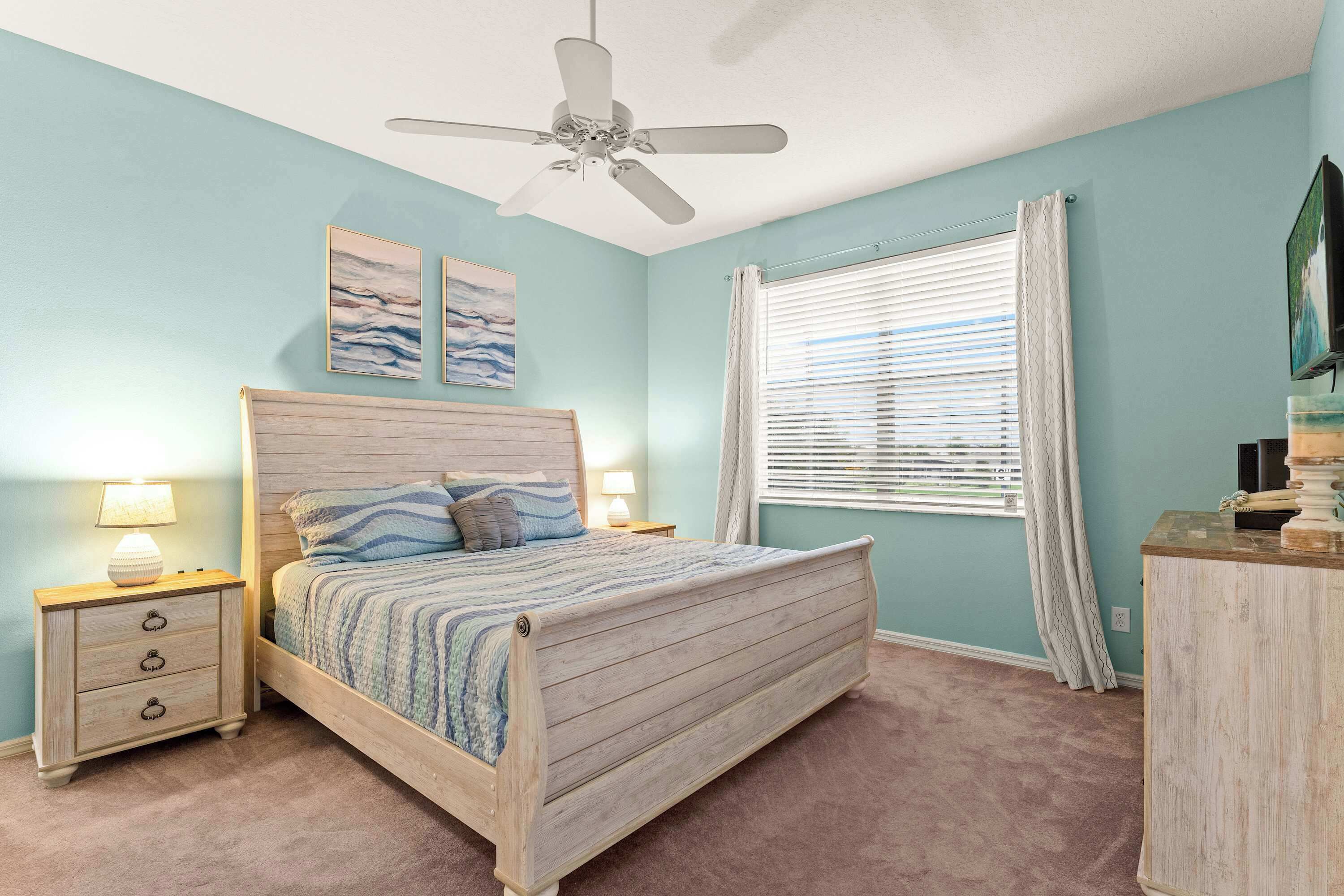 A bedroom with a bed, nightstands, a ceiling fan, a window with curtains, and artwork on the wall.