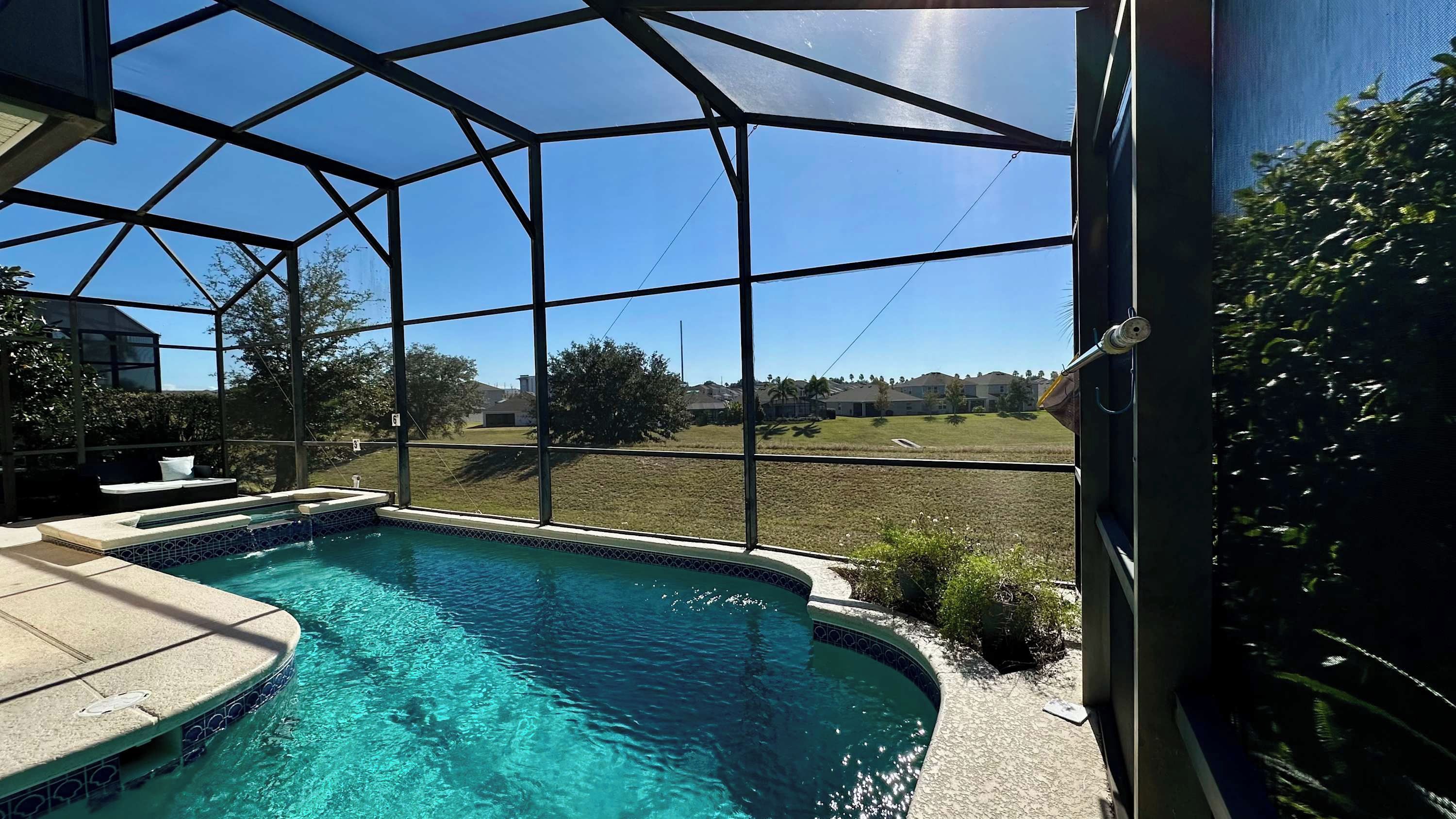 A swimming pool with a clear blue water, surrounded by a covered area with a metal frame and glass panels, offering a view of a grassy field and distant buildings.