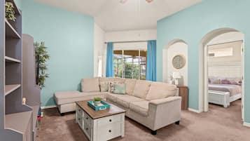 A living room with a beige sectional sofa, a white coffee table, and a bookshelf. There is a window with blue curtains, a ceiling fan, and a clock on the wall.