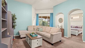 A living room with a beige sectional sofa, a white coffee table, and a bookshelf. There is a window with blue curtains, a ceiling fan, and a clock on the wall. - Poolside At Calabay - Home for 11 With Game Room, Pool & Hot Tub (Davenport)