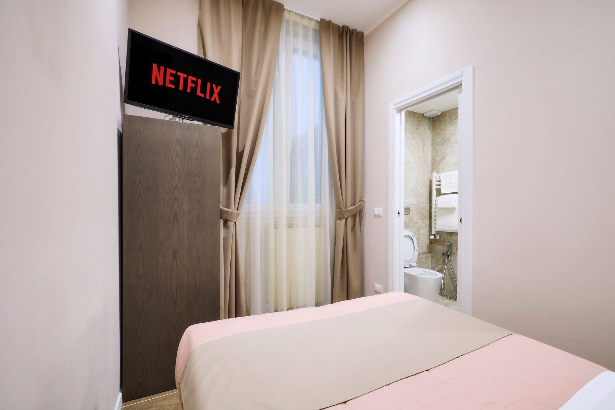 A hotel room with a bed, a TV displaying 'NETFLIX', a bathroom with a toilet and sink, and beige curtains.