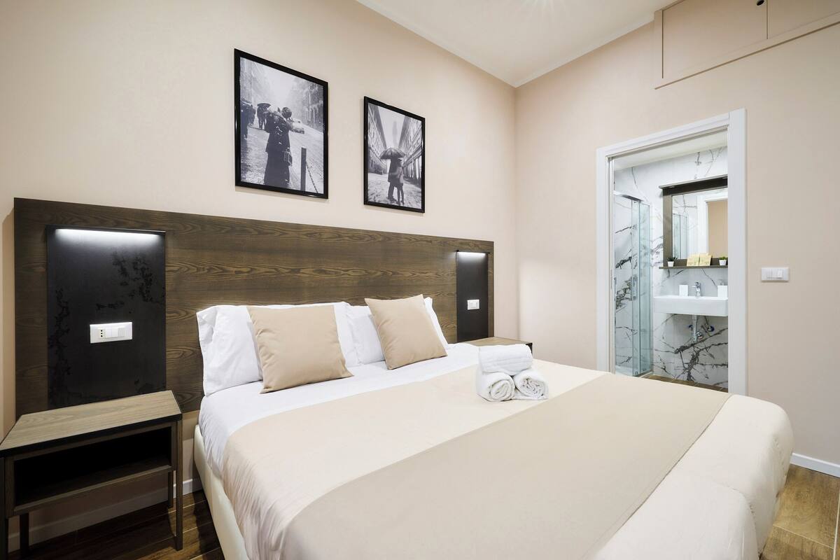 Habitación de hotel con una cama grande, cabecera de madera, dos cuadros enmarcados en la pared y un baño visible a través de una puerta abierta.