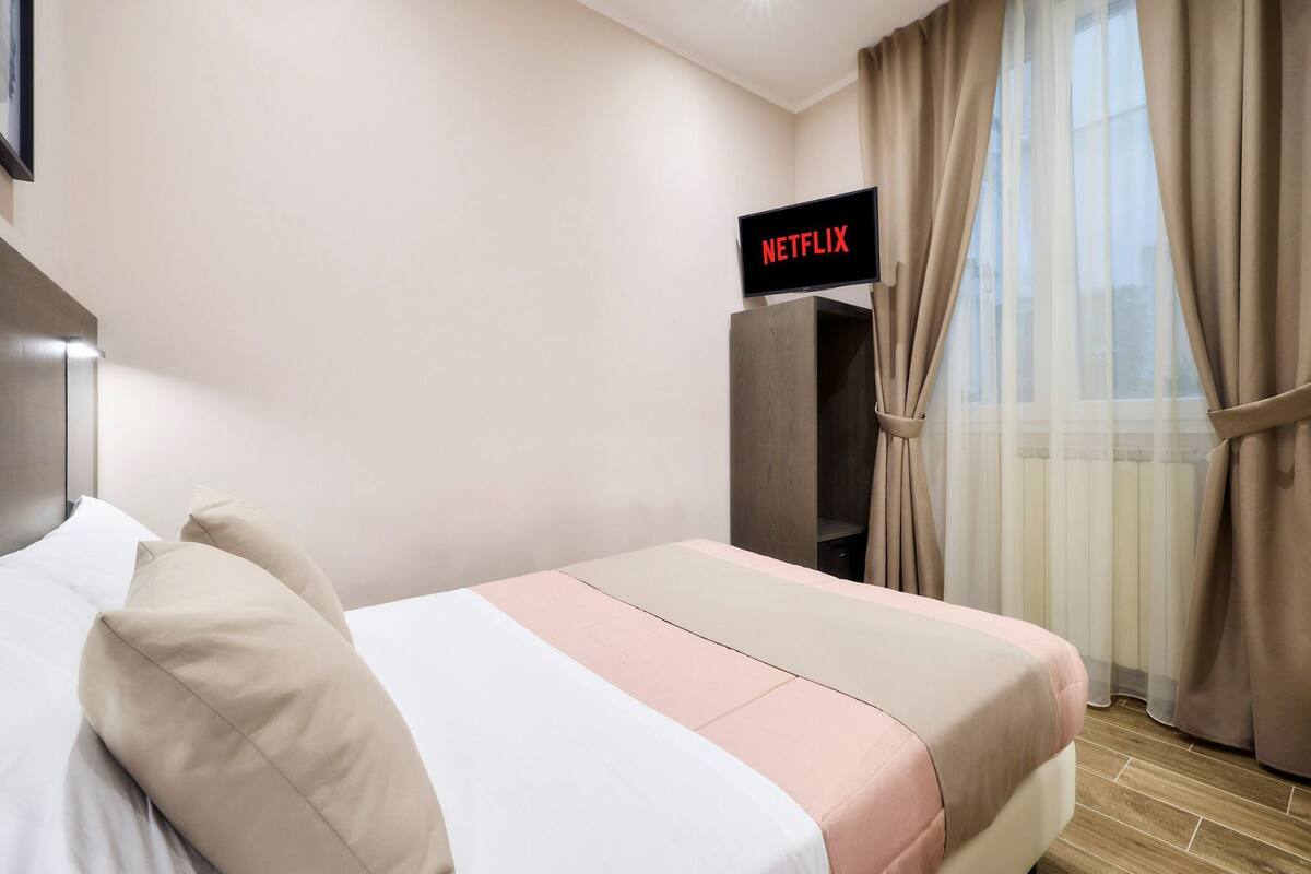 Una habitación de hotel con una cama, un televisor que muestra 'NETFLIX' y una ventana con cortinas.