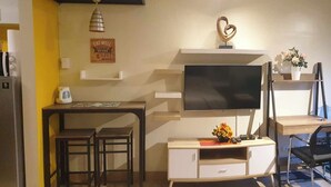 A compact living space with a TV mounted on a wall, a small dining table with chairs, a kitchenette with a microwave, and a fan near the window. - Spring Residences by VnB (Paranaque)
