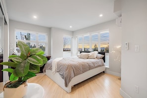 Een moderne slaapkamer met een groot bed, twee ramen met uitzicht op de stad, en een kamerplant.