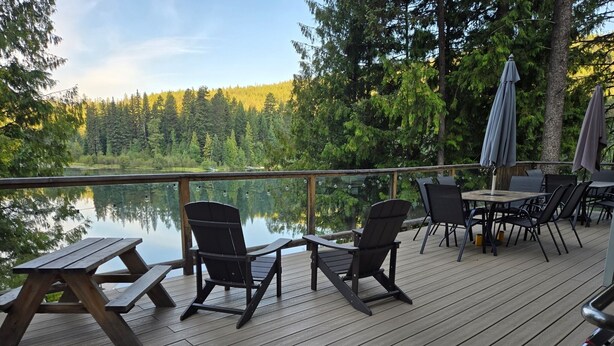 En träveranda med Adirondack-stolar, picknickbord och parasoller, med utsikt över en sjö.