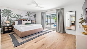 Ein Schlafzimmer mit einem großen Bett, einem Deckenventilator, einer Glastür zum Balkon und einem Wandgemälde mit tropischem Motiv.