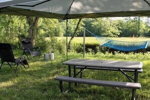 Un espace de pique-nique aménagé en plein air, avec une table et des chaises sous une tonnelle, un hamac et une zone herbeuse bordée d’arbres.