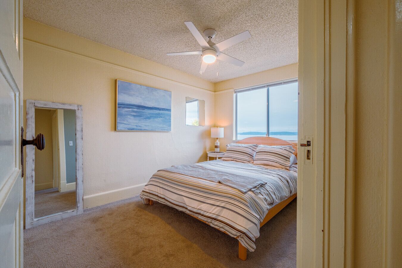 A bedroom with a bed, a ceiling fan, a window, and a painting on the wall.