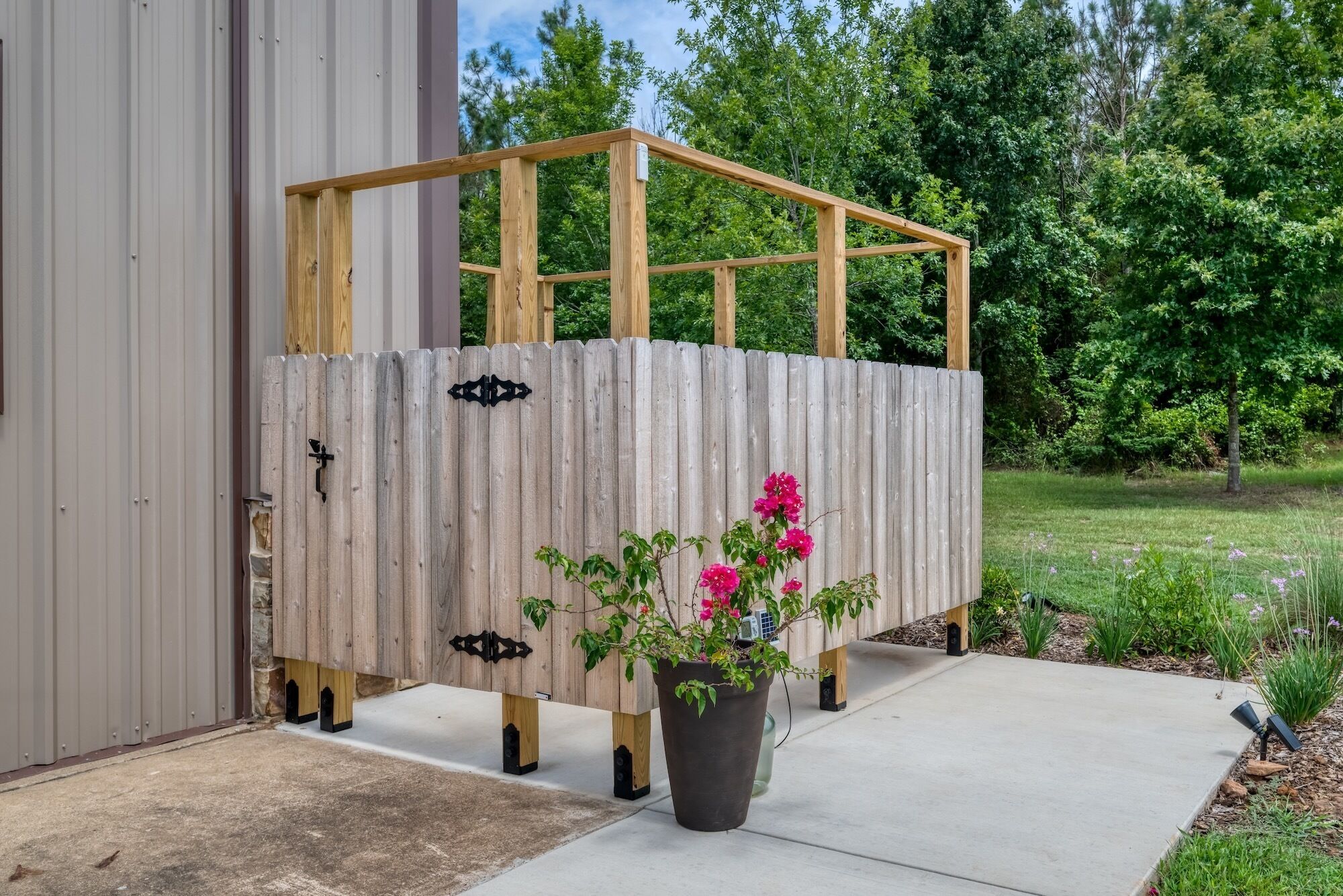 Une structure en bois avec une porte en métal, un pot de fleurs contenant des plantes aux fleurs roses et un chemin en béton.