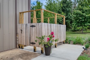 A wooden structure with a metal door, a potted plant with pink flowers, and a concrete pathway. - Private 300-Acre East Texas Retreat — Hot Tub, Stocked Pond, Total Seclusion (Jacksonville)
