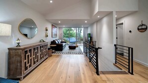 Amplia sala de estar con piso de madera, un espejo grande, un sofá y una mesa de centro. Se ve una escalera con barandal negro que conduce a otro nivel.