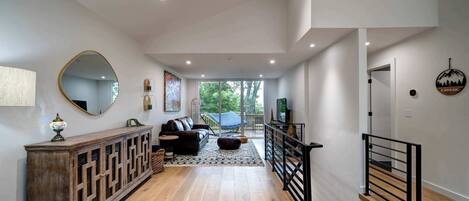 A spacious living area with a wooden floor, a large mirror, a sofa, and a coffee table. There is a staircase with a black railing leading to another level.