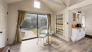 Une pièce spacieuse dotée d’une grande porte-fenêtre coulissante donnant sur un jardin, d’une table à plateau en verre avec des chaises, et d’un meuble blanc avec un plan de travail.