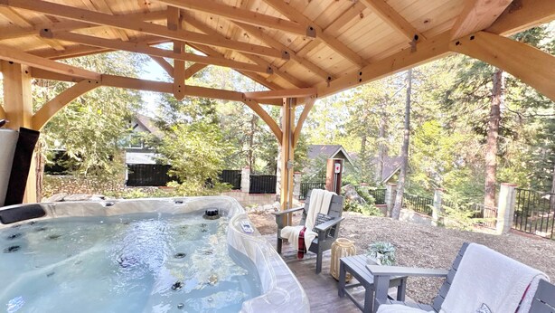 Un jacuzzi bajo una pérgola de madera con vistas a árboles y una casa al fondo.
