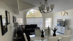 Een moderne woonkamer met een glazen eettafel, witte stoelen, een zwarte bank en een groot raam met jaloezieën.