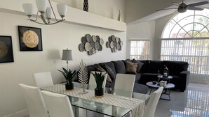 Een moderne woonkamer met een glazen eettafel, witte stoelen, een zwarte bank en een plafondventilator.