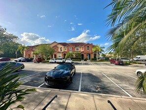 A parking lot with several cars, a building with a red facade, and palm trees. - Luxury Compass 3BR Townhouse Near Disney Parks 2 (Kissimmee)