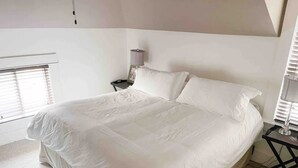 Una cama bien hecha en una habitación con ventana, una mesita de noche con lámpara y una pequeña mesa con otra lámpara.