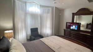 Une chambre d’hôtel avec un lit, un ventilateur de plafond, une télévision, une chaise et une commode.