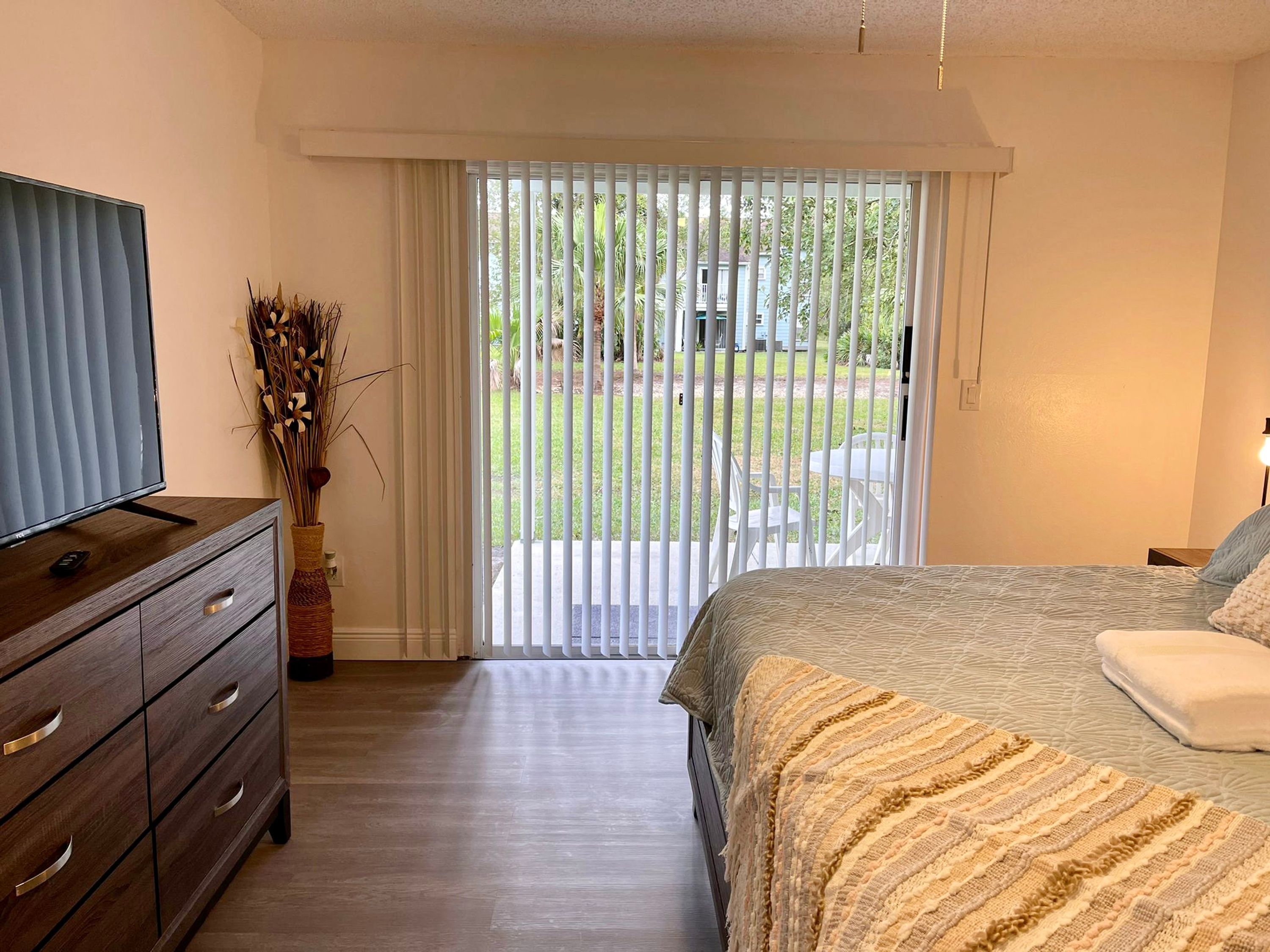 Un dormitorio con cama, cómoda, televisor y vista a un jardín por una puerta corrediza.