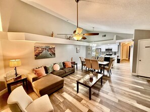 Un vaste salon comprenant un canapé, un pouf et une table basse, un coin repas avec des chaises, une cuisine à l’arrière-plan et un ventilateur de plafond.