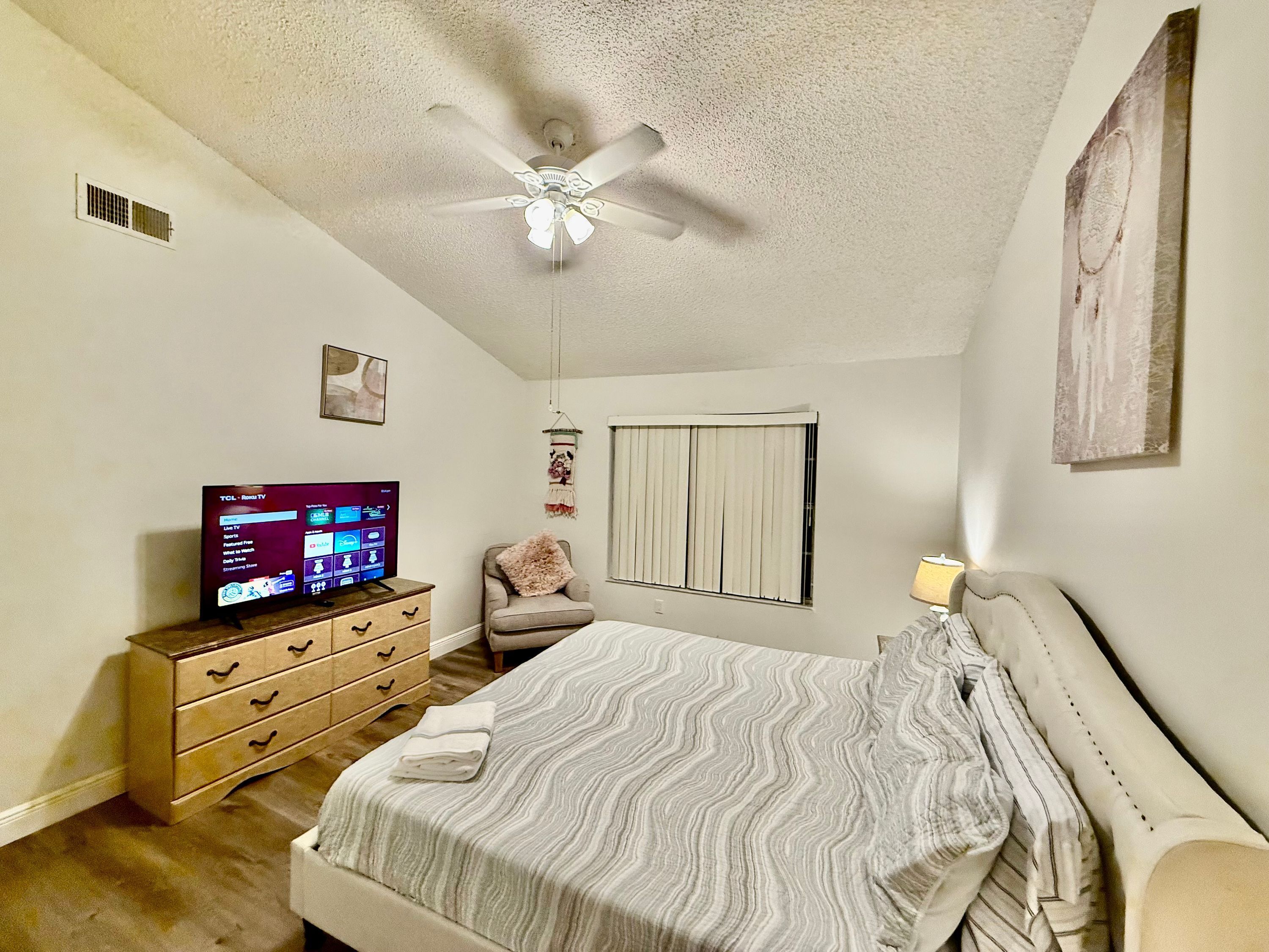 A bedroom with a bed, a dresser, a television, a ceiling fan, and a window with blinds.