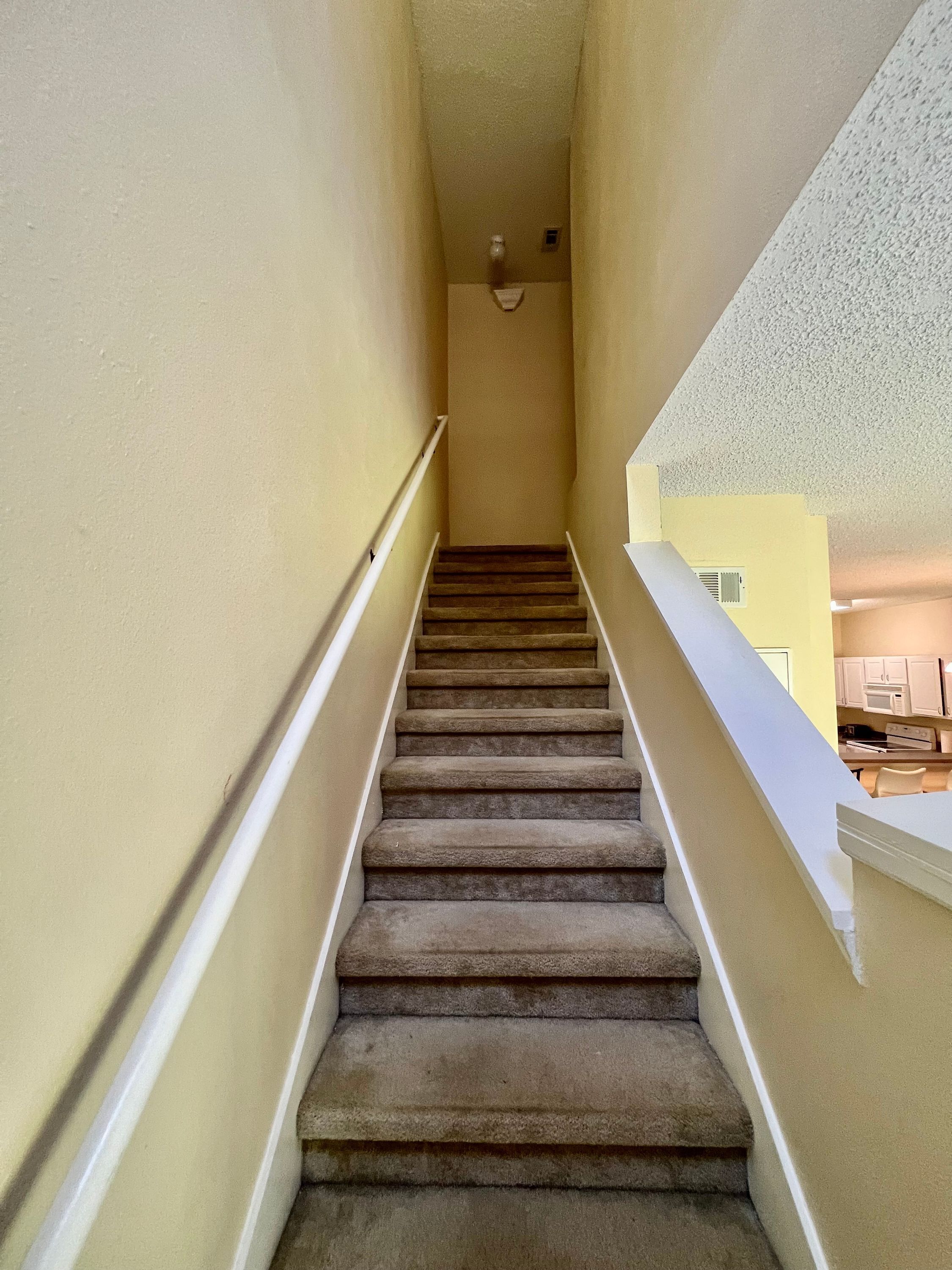 Una escalera empapelada con barandas blancas que conduce a un rellano, con una cocina visible al fondo.