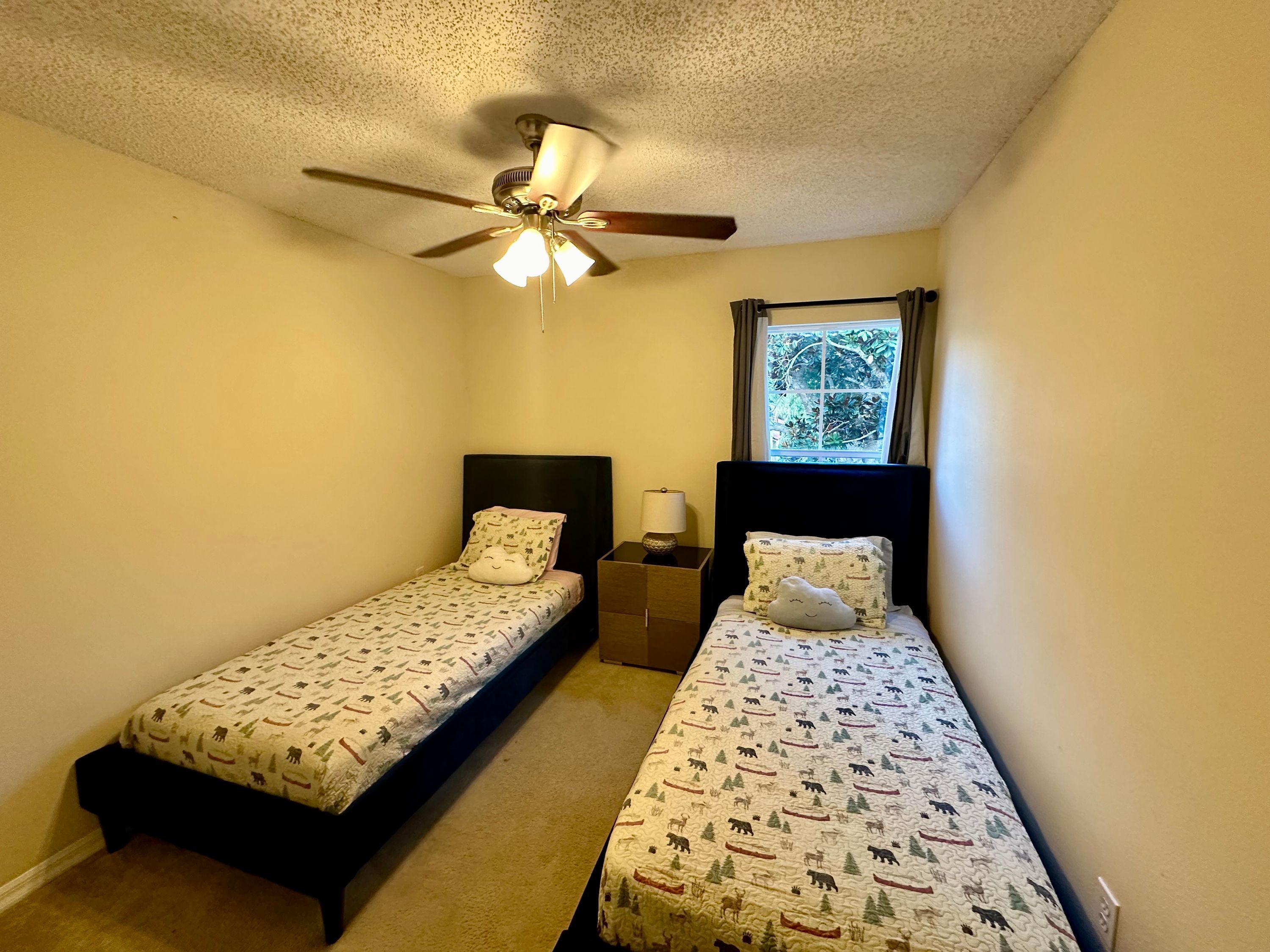 A small bedroom with two single beds, a ceiling fan, a window with curtains, and a nightstand with a lamp.