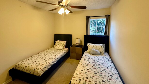 A small bedroom with two single beds, a ceiling fan, a window with curtains, and a nightstand with a lamp.