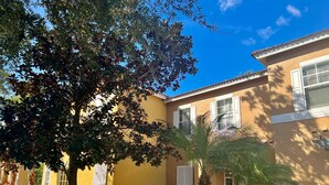 Un edificio amarillo con contraventanas blancas, rodeado de un jardín ajardinado con diversas plantas y una palmera.