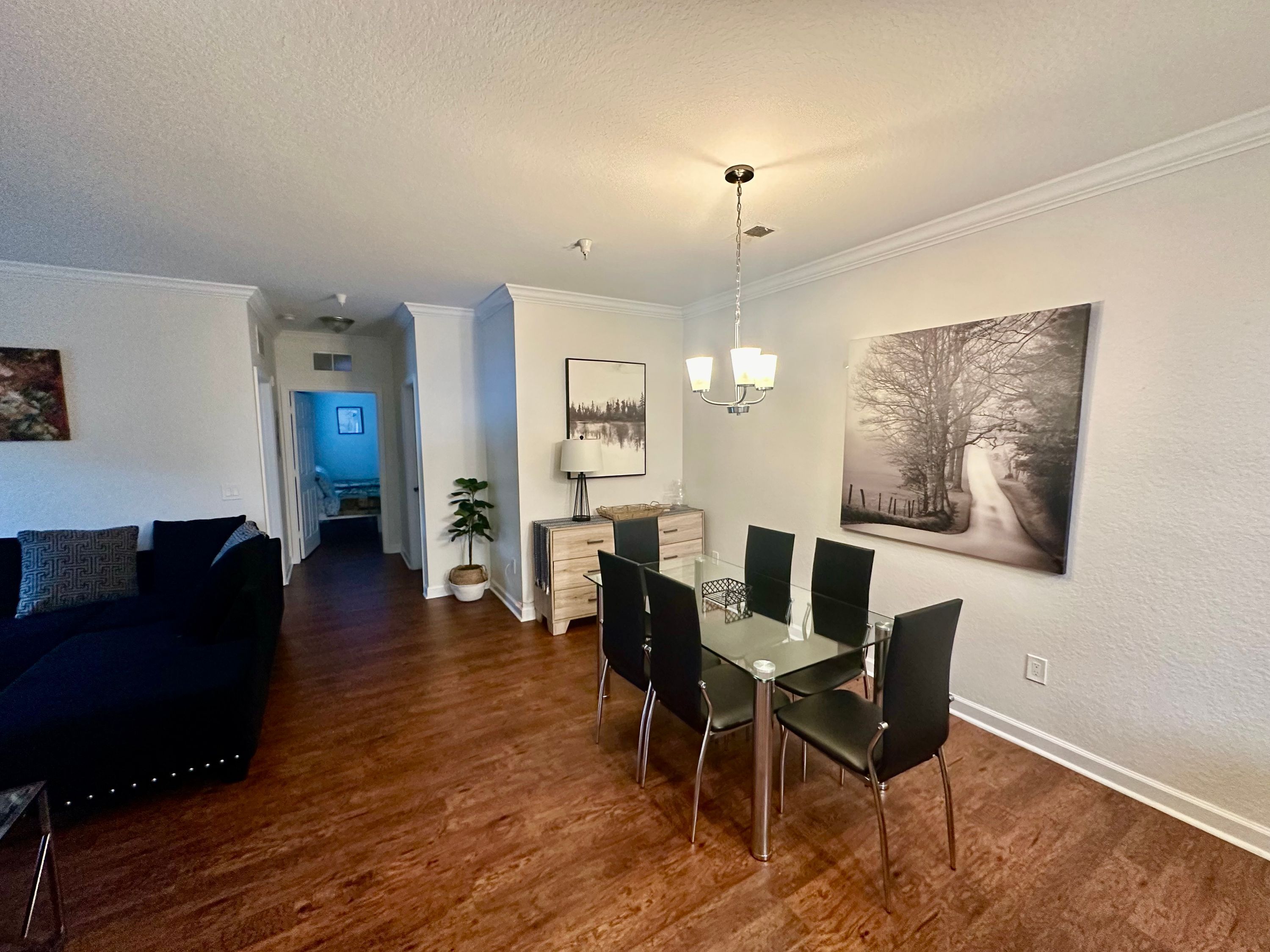 A modern living room with a dark-colored sofa, a dining area with a glass table and metal chairs, and a hallway leading to another room.