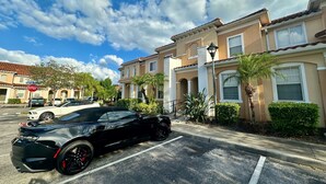 A black sports car parked in a residential street with Mediterranean-style houses, palm trees, and a clear blue sky. - Grand Resort 3BR Compass Townhouse Near Disney 1 (Kissimmee)