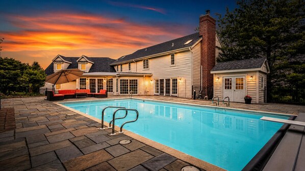 A swimming pool with a patio area, a covered seating area, and a house with a brick chimney.