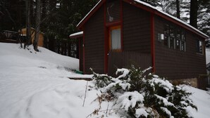 Une cabane en bois avec une porte rouge et des fenêtres, entourée d’un paysage enneigé et d’arbres.